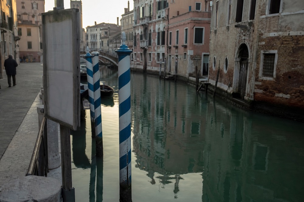 Striped mooring poles standing in green water beside a stone quay, with a laminated notice board fixed to a wooden post