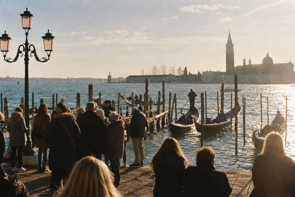 Crowd of visitors waiting on a stone waterfront beside moored gondolas, open lagoon water and distant church tower beyond