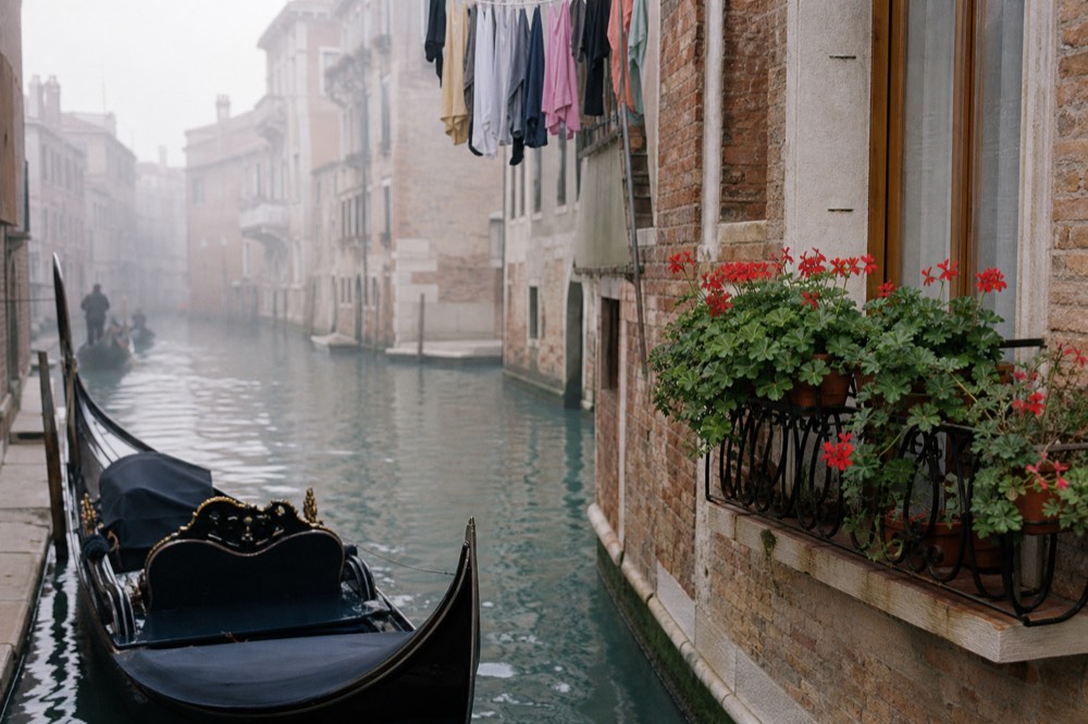 A single gondola resting on flat water in a residential canal with brick walls, potted geraniums on a balcony and hanging laundry above