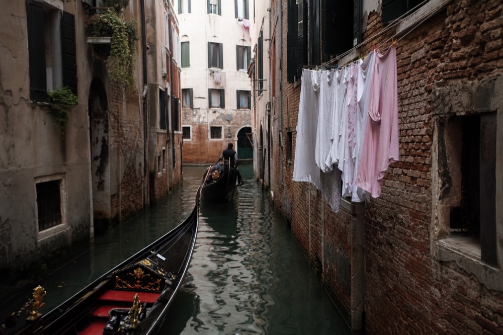 A narrow canal barely wider than a boat, with laundry lines strung between shuttered windows and green water reflecting the walls
