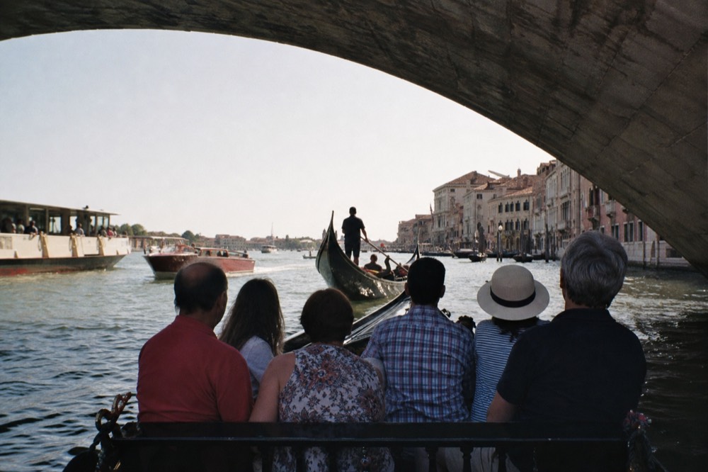 Six passengers sharing a bench in a gondola on wide open water with several other boats visible around them in flat afternoon light