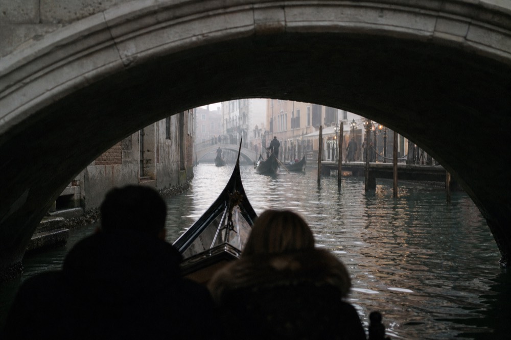 A couple's silhouetted shoulders in the front seat of a gondola passing beneath a low stone bridge, water lit from the far side